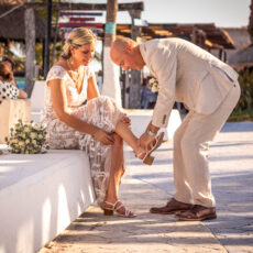 A groom helps his new bride remove her shoes to go for a walk on the beach together in Puerto Morelos.