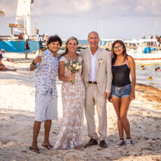 The bride and groom celebrate on the beach with one male and one female friend. In the background, many people are present enjoying the beach, the iconic leaning lighthouse and the pier.