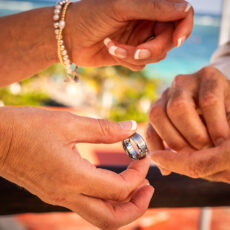 Photo of the hands of a bride and groom, where the bride is about to place the groom's wedding ring on his finger.