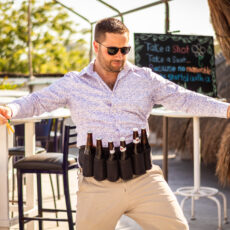 A young man wearing a special beer holster belt full of 6 beer bottles, with another opened beer in his hand.