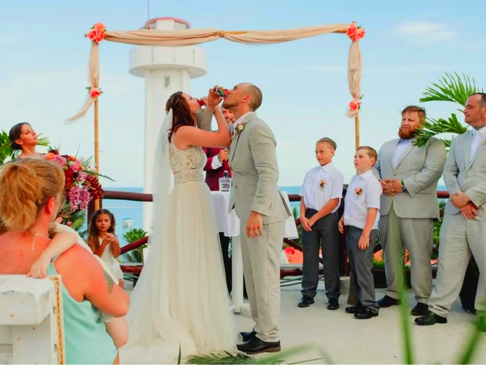 A newly married couple stands on the rooftop of La Sirena Puerto Morelos overlooking the iconic leaning lighthouse in front of all of their family and friends while sharing a first cocktail as a married couple.